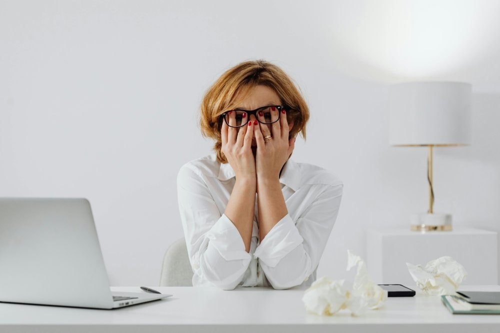 A frustrated woman covering her face with her hands at a desk in front of a laptop