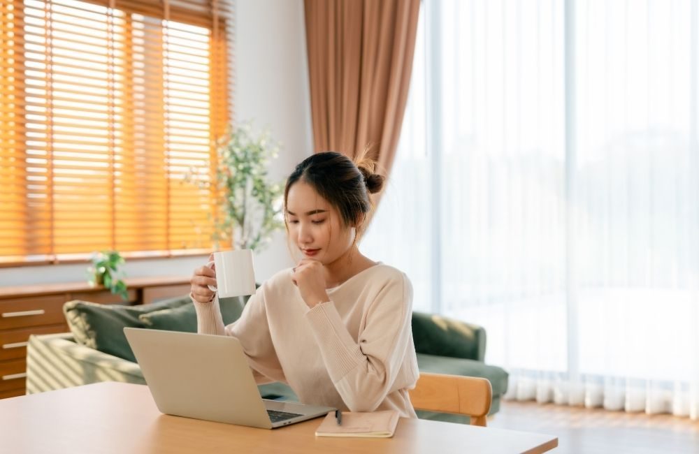 A woman smiling at her laptop in a sunny living room, holding a mug