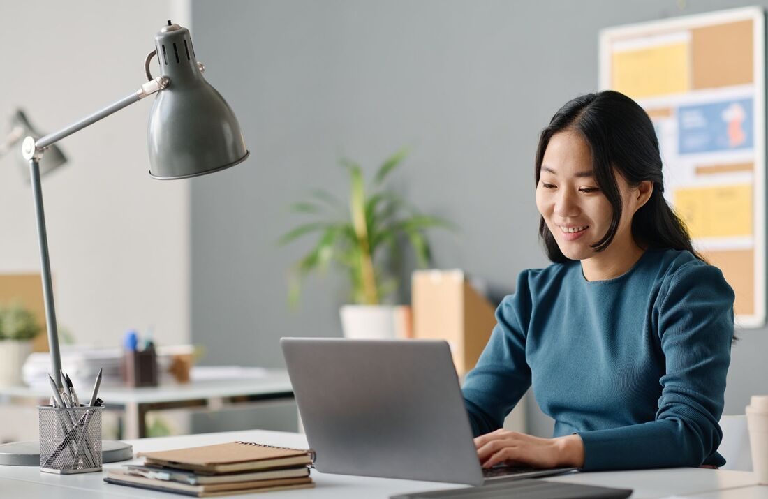 A woman smiling as she works on her laptop at a bright home-office desk
