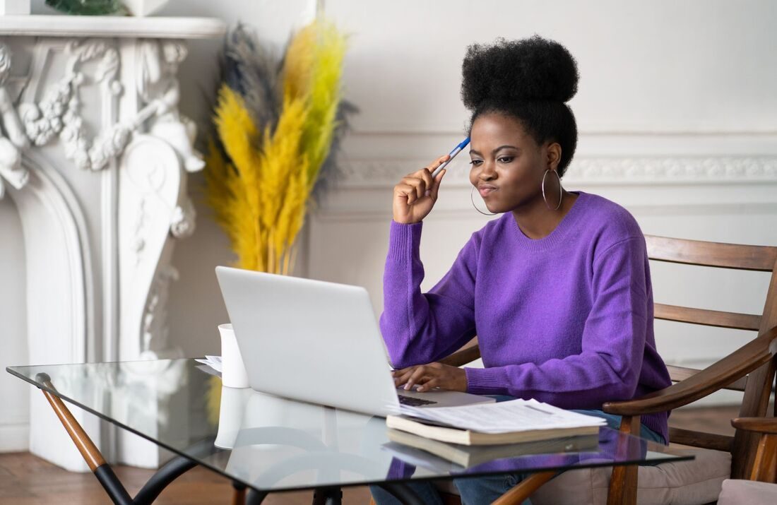 A woman in a purple sweater thinking as she works on a laptop at a glass desk