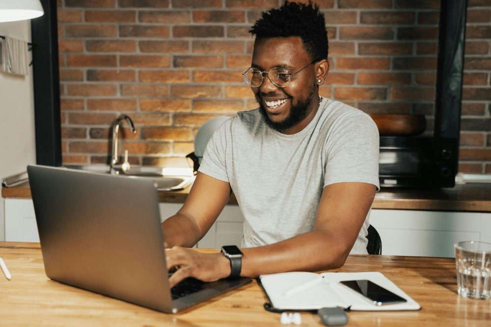 A smiling man working on a laptop at a wooden table in front of a brick wall