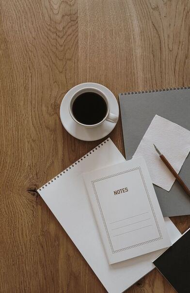 A cup of black coffee, a notes booklet, and a pencil on a warm wooden desk