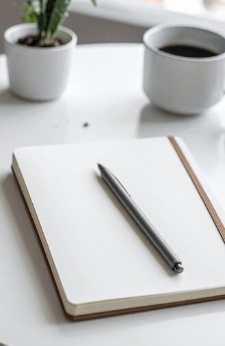 An open notebook and a pen beside a small plant and a cup of coffee on a white table