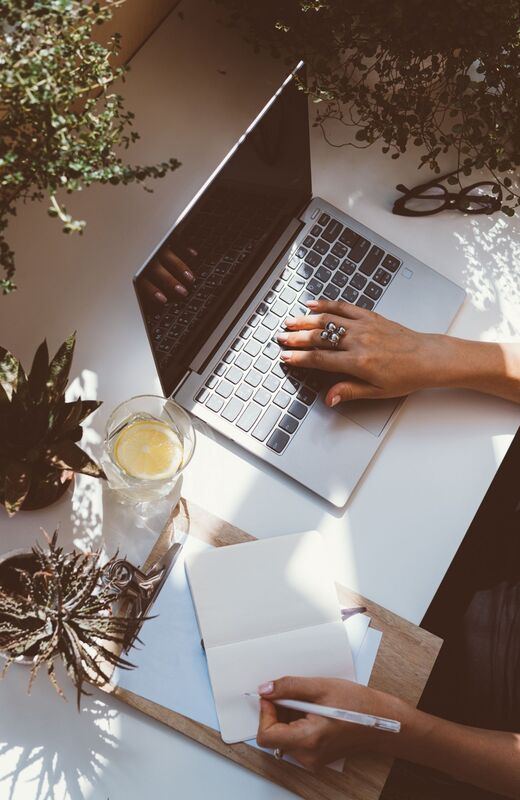 Hands typing on a laptop at a white desk with dried flowers and a glass of lemon water