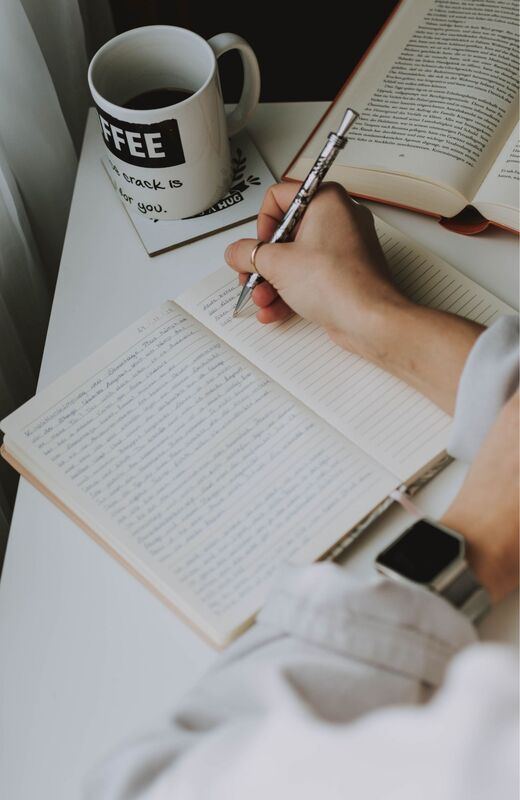 A hand writing in a journal beside a coffee mug and an open book